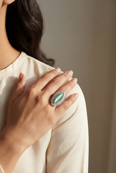 Close-up of a hand wearing an ornate ring with a green gemstone, against a neutral background.