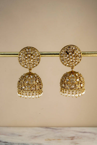 Close-up of gold-toned earrings with detailed American Diamond embellishments and pearl accents, displayed on a jewelry stand.