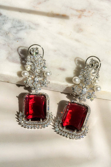 Flat lay of Aayat Statement Earrings in ruby red featuring a bold gemstone, floral pavé design, and pearl detailing. Rhodium-plated danglers.