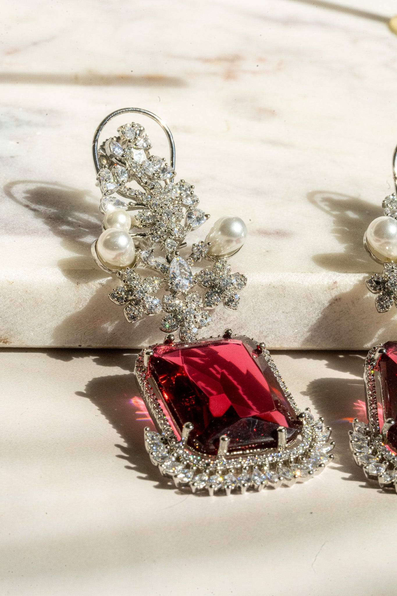 Angled close-up of ruby red Aayat Statement Earrings with radiant gemstone, pavé halo, and pearl floral accents. Rhodium-plated finish.
