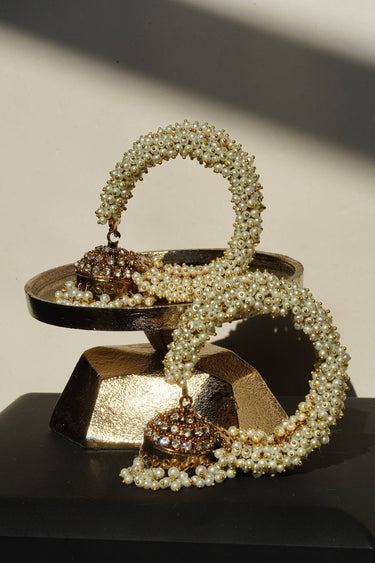 A pair of Aliya Jhumka Bangle Bracelets in antique gold with faux pearl beadwork, coiled on a tiered dark metal display prop.