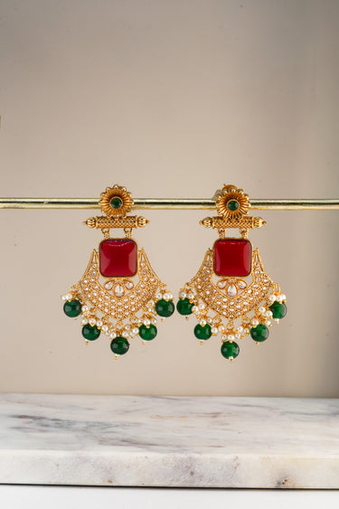 A symmetrical studio front view of the Kavya statement earrings featuring a vibrant ruby red centerpiece stone and emerald green cascading glass drops.