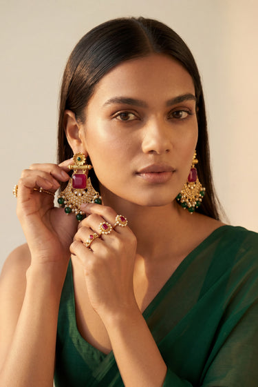 Close-up front portrait of a model adjusting the Kavya gold-plated crescent statement earrings featuring ruby centerpieces and green glass drops.