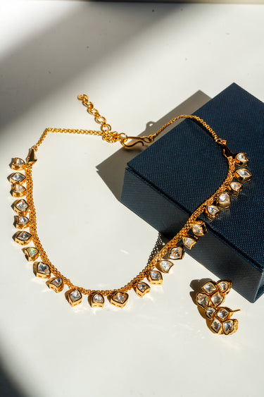 A full retail flat lay shot of the gold-plated Kiara heart-cut Kundan strand necklace and one matching stud earring displayed next to a dark navy premium jewelry gift box.