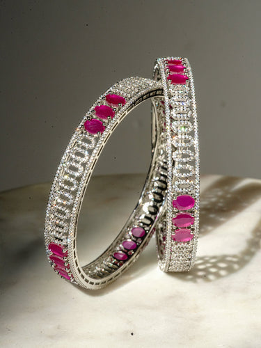 An upright studio product display of the pair of rhodium plated Kyra statement bangles featuring brilliant white pave AD stones and ruby pink gemstone sections.