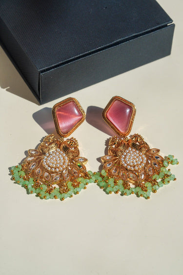 A top-down studio shot of the Mira statement earrings featuring blush pink rhombus top stones and mint green bead droplets next to a black box