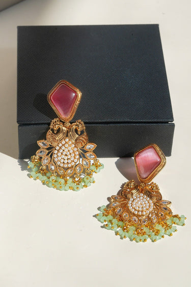 A detailed presentation shot of the pink and mint green Mira peacock drop earrings arranged flat on a white surface with a luxury black jewelry box packaging background.