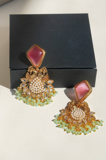 A detailed presentation shot of the pink and mint green Mira peacock drop earrings arranged flat on a white surface with a luxury black jewelry box packaging background.