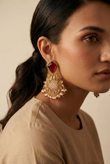 A detailed side view portrait of a model showcasing the deep ruby red variant of the Mira traditional peacock earrings with matching red glass droplets.