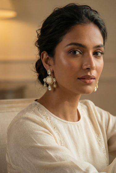 A close up three quarter lifestyle portrait of a model styling the mehndi gold plated Zoe geometric drop earrings with a cream colored outfit.