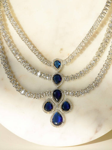 Niara Triple Layer Diamante Accent AD Necklace Set in gold or rhodium-plated silver finish with color options of silver & sapphire blue, silver & green, gold & green, and silver & ruby, featuring triple-layered chain links and matching statement earrings.