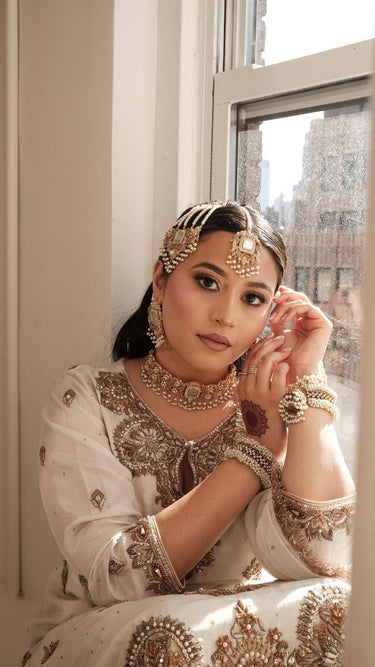 Woman in Pakistani traditional attire with paksitani bridal jewelry, leaning against a window.