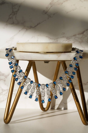 Aagya AD Necklace Set – Rhodium Plated in blue with teardrop stones and CZ detailing, displayed on a marble and gold stand.