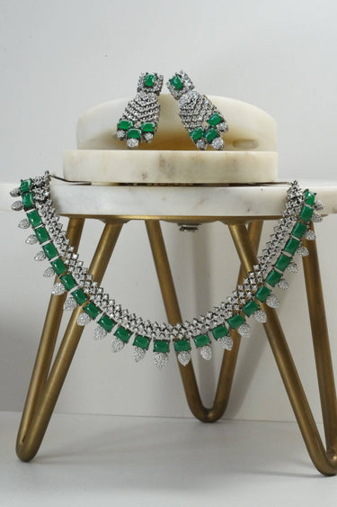 Front view of green Anjam Black Plated AD Necklace Set with matching earrings, styled on a marble platform with gold legs.