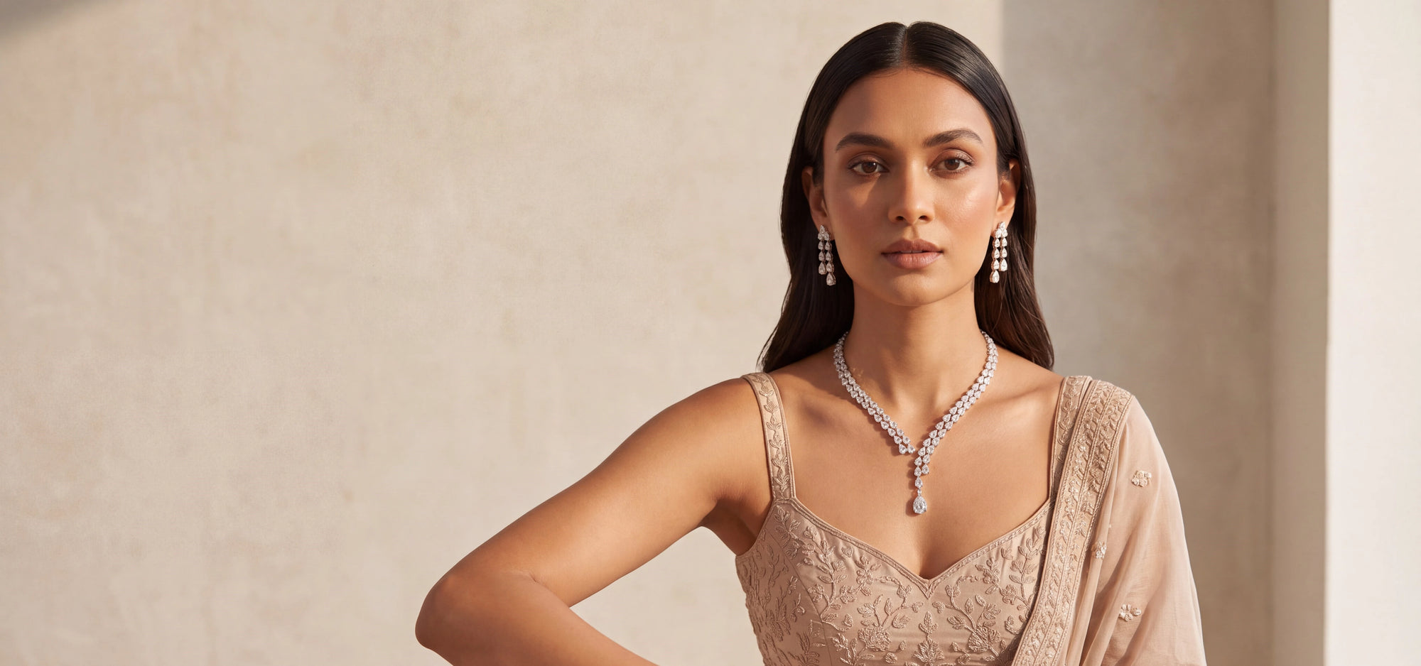 A woman modeling a delicate AD diamante style necklace with a pear-cut center stone and matching drop earrings from Inaury.com, wearing a beige embroidered dress against a textured beige wall.