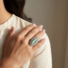 Close-up of a hand wearing an ornate ring with a green gemstone, against a neutral background.