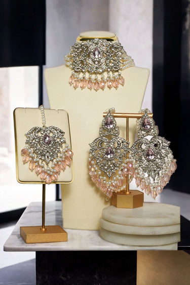 Noreen Choker Necklace Set - Elegant Pakistani Bridal Jewels in silver with pink teardrop stones, displayed with earrings and tikka on stand.