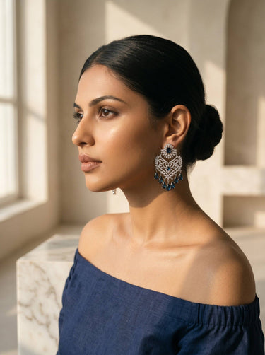 Woman wearing a blue off-shoulder top and medium size decorative Indian Ad earrings in rhodium plating with sapphire blue teardrop stones, in a softly lit room. 