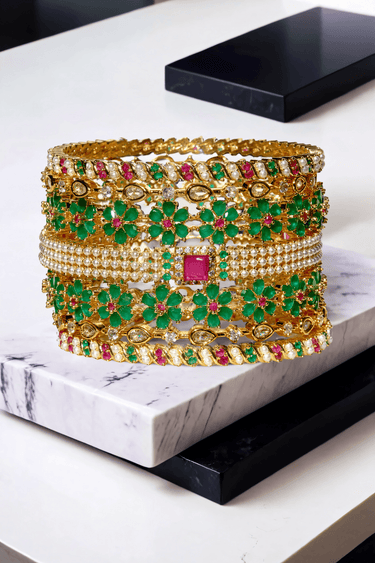 Arika gold-plated floral bangle set with emerald and ruby accents displayed on marble. Elegant 7-piece jewelry for weddings and festive wear.