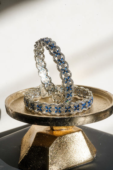Rhodium plated Indian bangles with sapphire blue and white stones on a metallic stand against a white background
