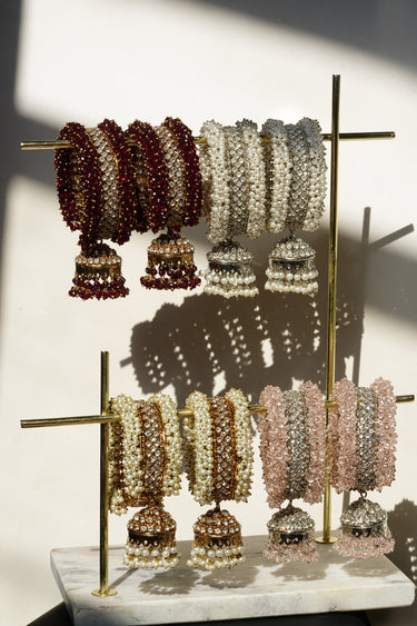 The Aliya Jhumka Bangle Bracelet in antique gold with faux pearl beadwork, shown on a wooden display rack alongside other colorways.