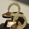 A pair of Aliya Jhumka Bangle Bracelets in antique gold with faux pearl beadwork, coiled on a tiered dark metal display prop.