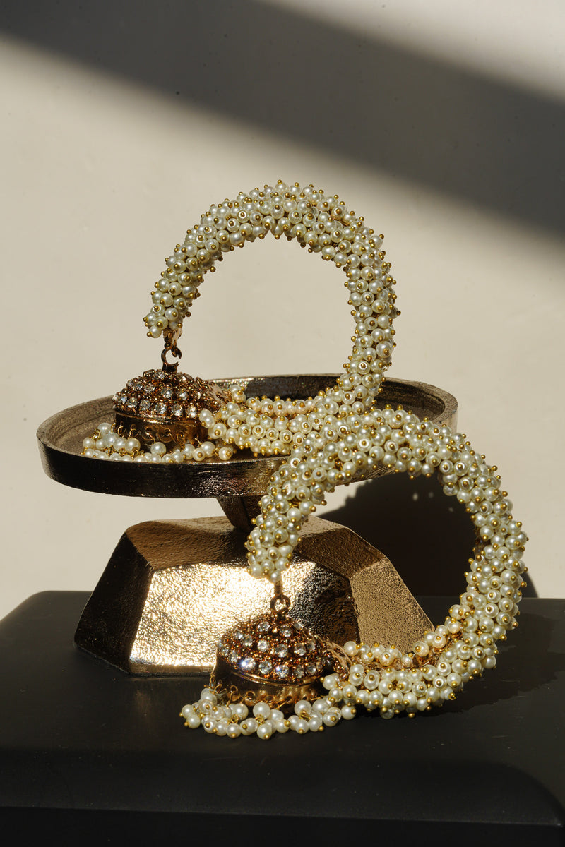 A pair of Aliya Jhumka Bangle Bracelets in antique gold with faux pearl beadwork, coiled on a tiered dark metal display prop.