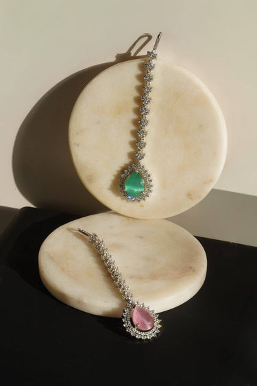 Two Amore rhodium-plated Maang Tikkas showcasing the emerald green and light pink teardrop crystal variants displayed on marble stones.