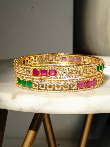 A horizontal studio layout of the gold plated Kyra statement bangles stacked to showcase the alternating sections of emerald green and ruby pink square cut stones.