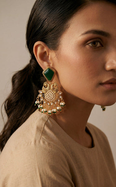 A close up side profile portrait of a model styling the emerald green variant of the gold plated Mira peacock statement drop earrings.