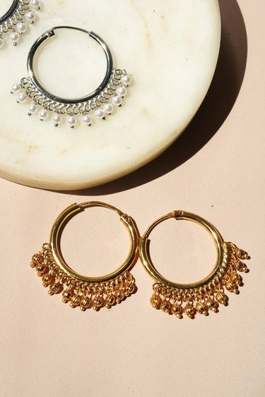 A crisp product layout of the pair of gold plated Sia ghungroo hoop earrings resting on a cream marble display disk.