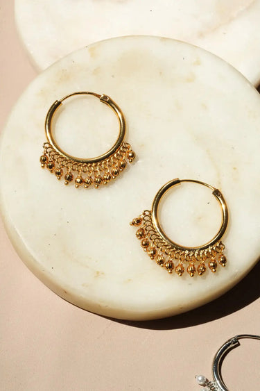 An extreme close up photograph of a single gold plated Sia hoop earring highlighting the detailed ghungroo bead fringe.