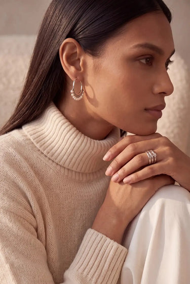 A side profile lifestyle portrait of a model wearing the rhodium plated Sia hoop earrings with pearl bead fringe accents paired with a cream turtleneck sweater.