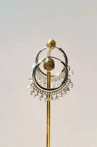 A vertical studio shot of the rhodium plated Sia pearl hoop earrings hanging on a polished brass earring stand.