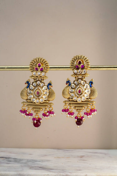 Inaury Adrita Kundan Meenakari Peacock Drop Earrings pair on gold bar stand — full view of both earrings showing four-tier temple jewelry silhouette with peacock motif and cascading ruby and pearl drops