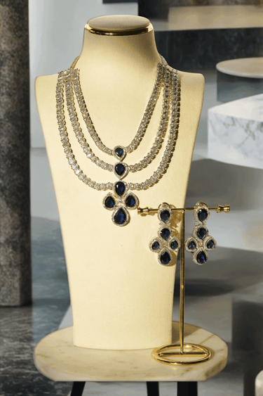 Niara Triple Layer Diamante Accent AD Necklace Set in gold or rhodium-plated silver finish with color options of silver & sapphire blue, silver & green, gold & green, and silver & ruby, featuring triple-layered chain links and matching statement earrings.