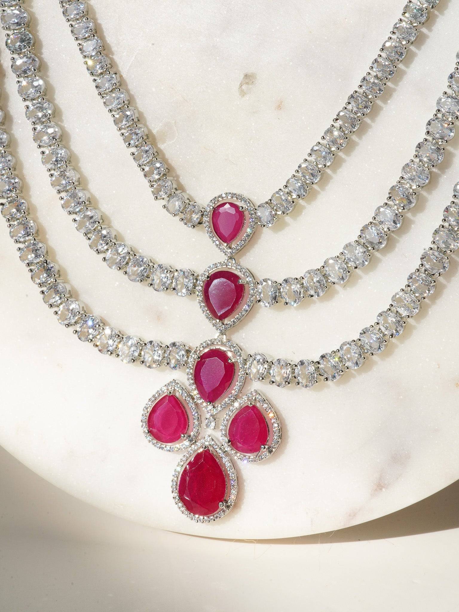 Niara Triple Layer Diamante Accent AD Necklace Set in gold or rhodium-plated silver finish with color options of silver & sapphire blue, silver & green, gold & green, and silver & ruby, featuring triple-layered chain links and matching statement earrings.