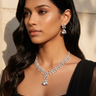 A woman with long, wavy black hair models a sparkling silver-tone diamond necklace and matching drop earrings against a warm beige stone wall background. Indian statement jewelry set by Inaury