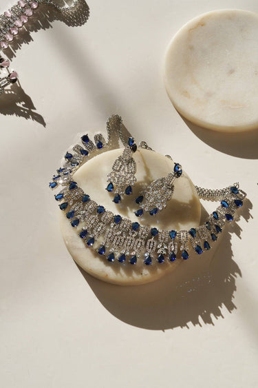 Aagya AD Necklace Set – Rhodium Plated in blue, laid out on marble with earrings, showcasing South Asian jewelry elegance.