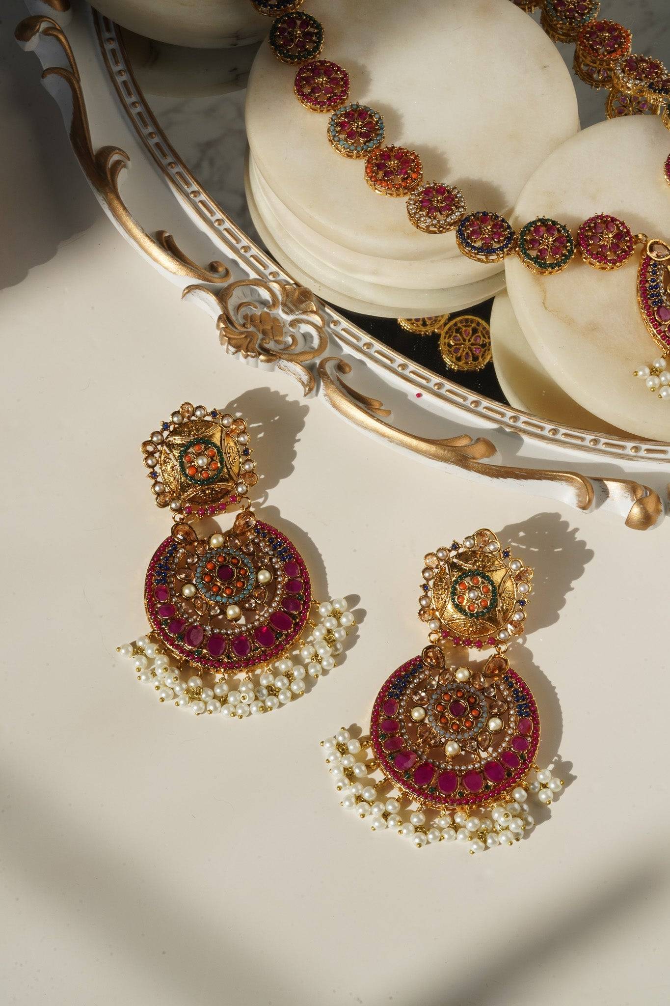 Pair of Adiva Statement Earrings with gold plating, pink and green stones, and pearl detailing, placed beside necklace.