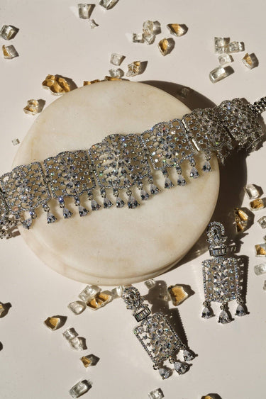 Flat lay of Nafisha Silver & AD Stone Choker Set with earrings on marble, surrounded by decorative crystals for a luxury jewelry display.