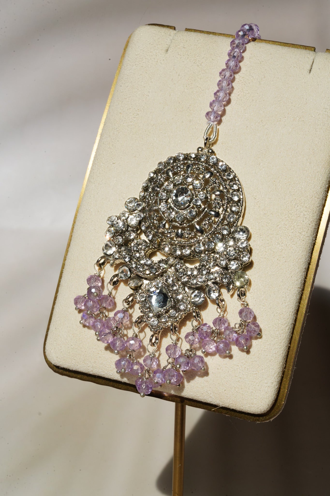 Maang tikka from Namar Choker Necklace Set with lavender beads and CZ stones displayed on gold stand.